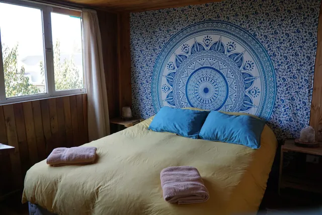 Habitación luminosa con cama matrimonial, tapiz de mandala decorativo azul y detalles en madera que crean un ambiente cálido y relajante — Villarrica — Hostal Micelio, Villarrica.