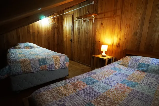 Habitación revestida en madera, con dos camas cubiertas por coloridas mantas patchwork y una lámpara cálida que realza el ambiente acogedor y tranquilo — Huerquehue — Hostal Micelio, Villarrica.