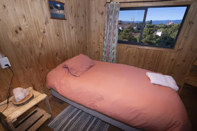 Habitación con revestimiento de madera clara, cama individual con ropa de cama color salmón y un ventanal que ofrece vista al paisaje y al cielo abierto — Pumalín — Hostal Micelio, Villarrica.