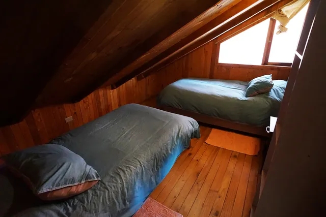 Habitación de madera con dos camas individuales y luz natural que entra por la ventana, creando un ambiente cálido y tranquilo — Nahuelbuta — Hostal Micelio, Villarrica.