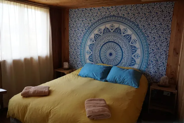 Cálida habitación con cama matrimonial, tapiz azul decorativo y cortinas claras que dejan pasar la luz del día, creando un ambiente relajante y armonioso — Villarrica — Hostal Micelio, Villarrica.