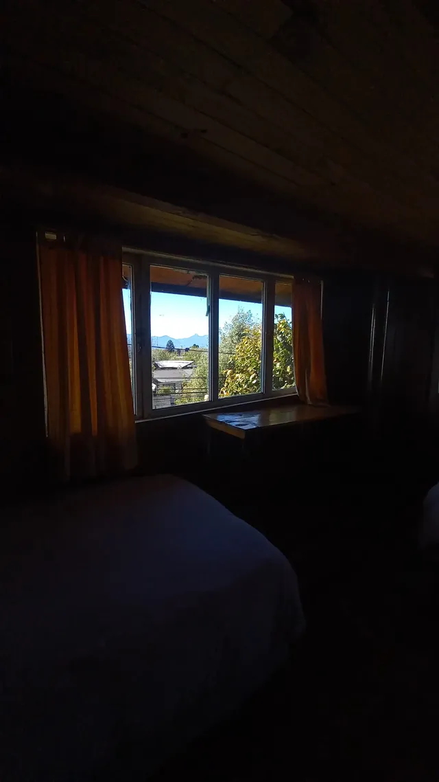 Habitación revestida en madera con grandes ventanales y cortinas doradas, donde la luz natural resalta la calidez del espacio y revela una vista tranquila hacia el lago Villarrica — Conguillío — Hostal Micelio, Villarrica.