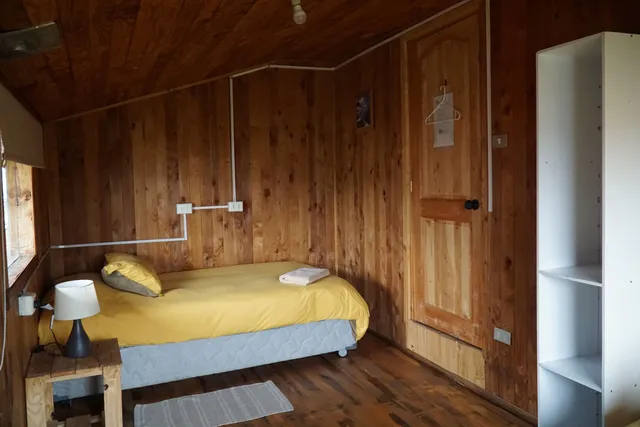 Cama con almohadas suaves y estante blanco, iluminado por luz natural, en un entorno tranquilo que refleja la esencia de Villarrica - Torres del Paine - Hostal Micelio Villarrica