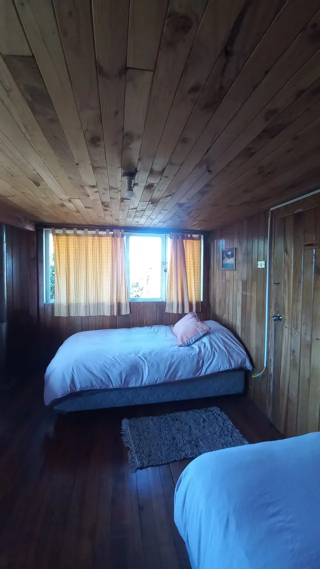 Habitación de estilo rústico con paredes y techo de madera, cortinas color miel que filtran la luz del sol, y una alfombra tejida junto a camas confortables, creando un ambiente cálido y tranquilo — Conguillío — Hostal Micelio, Villarrica.