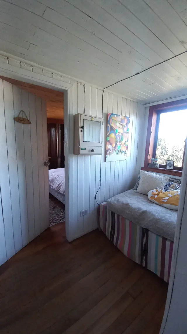 Sala común luminosa junto a la entrada de la habitación, con muros blancos y piso de madera. Un rincón acogedor con diván colorido, arte local y vista al volcán Villarrica — Conguillío — Hostal Micelio, Villarrica.